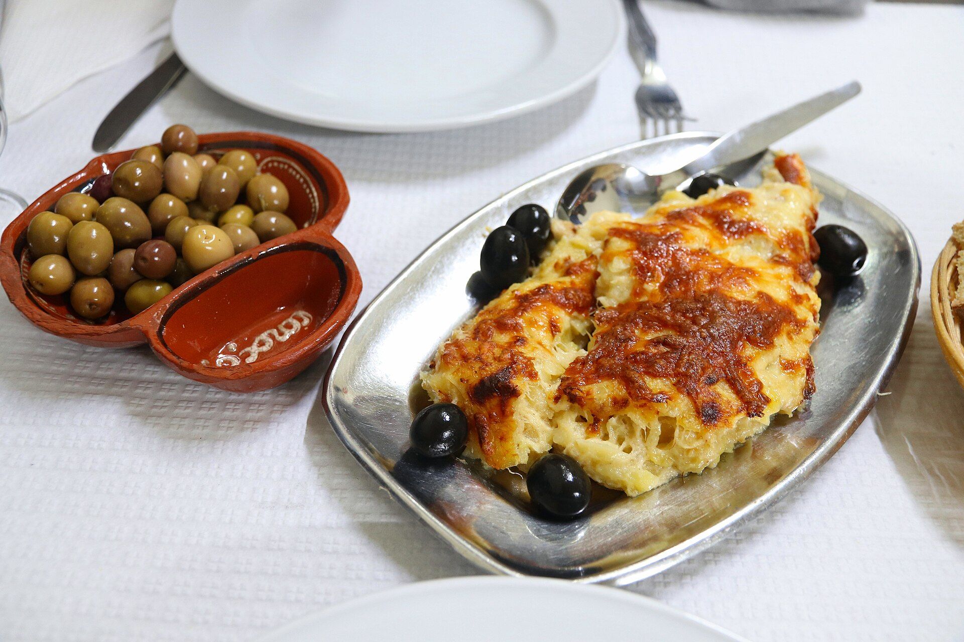 Travessa de bacalhau com natas servida num restaurante, com um prato de azeitonas ao lado