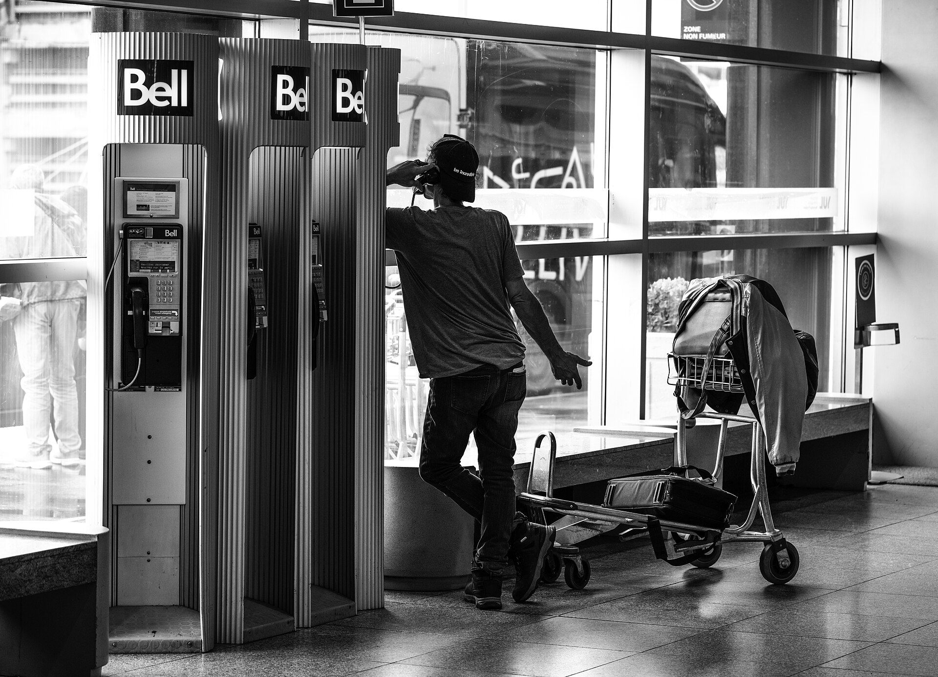 Homem encostado a uma fila de cabines telefónicas num aeroporto, em fotografia a preto e branco