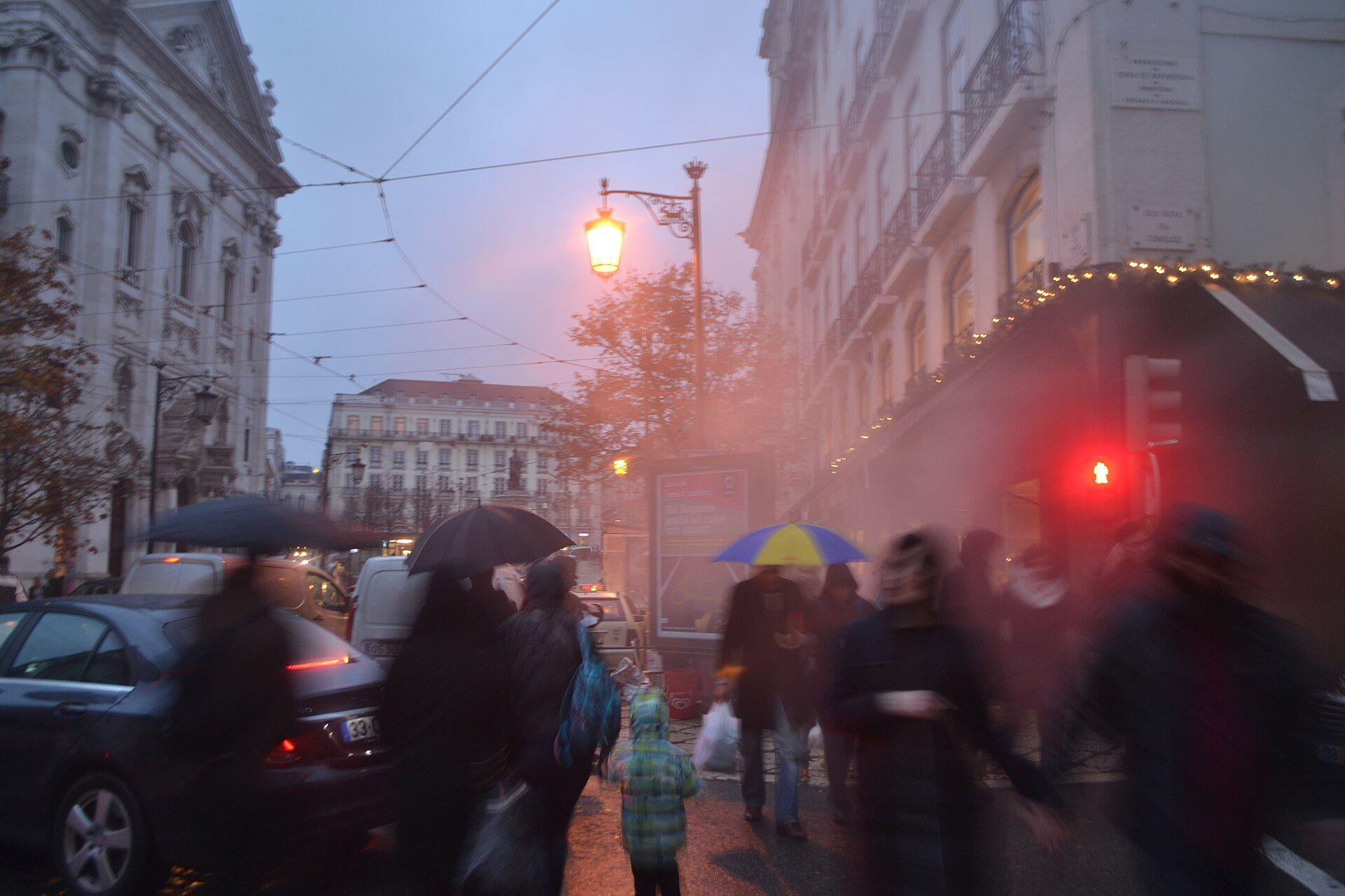 Rua de Lisboa num fim de tarde de chuva, com pessoas de guarda-chuva e um semáforo vermelho