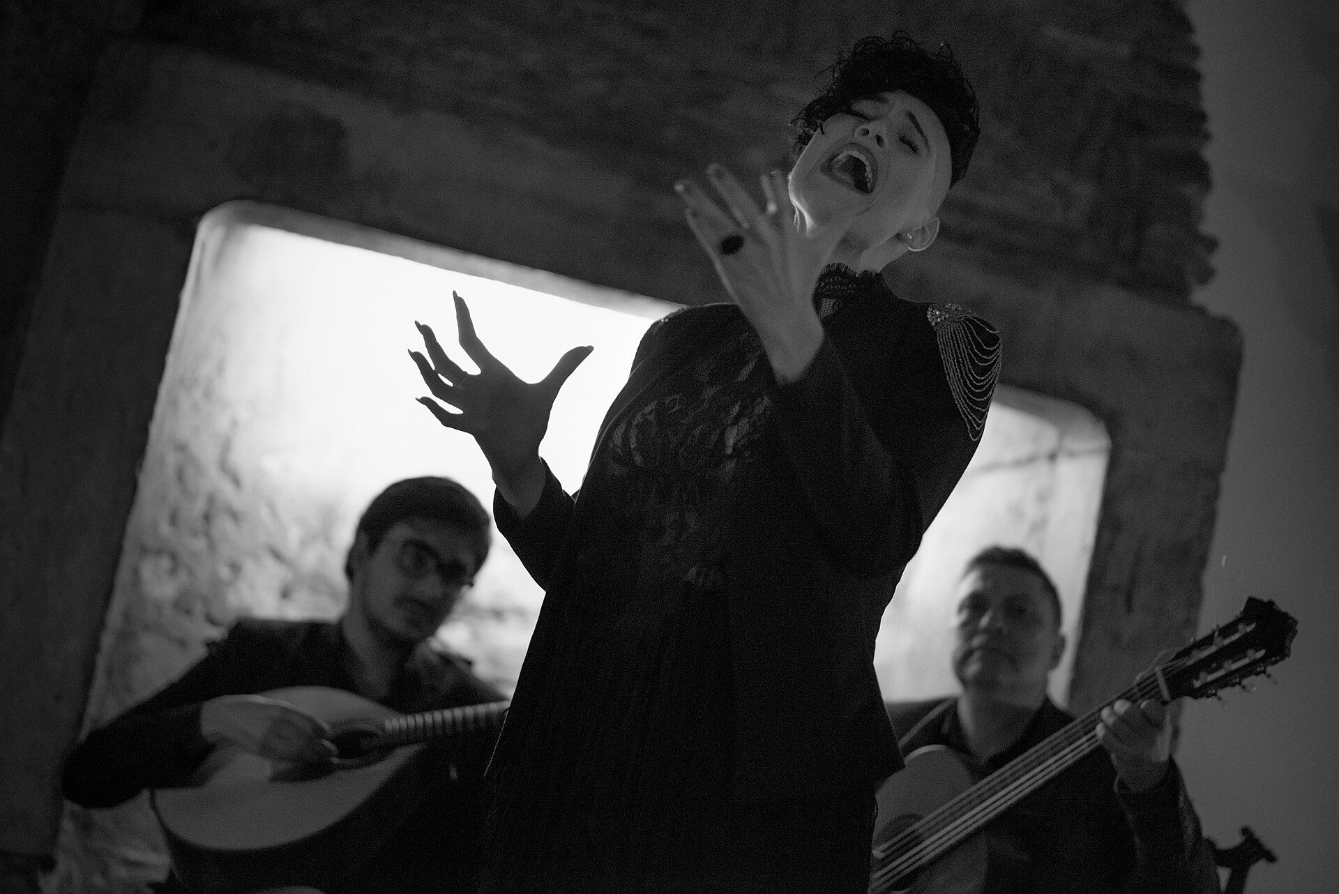 Fadista a cantar de pé, acompanhada por dois guitarristas sentados, em fotografia a preto e branco