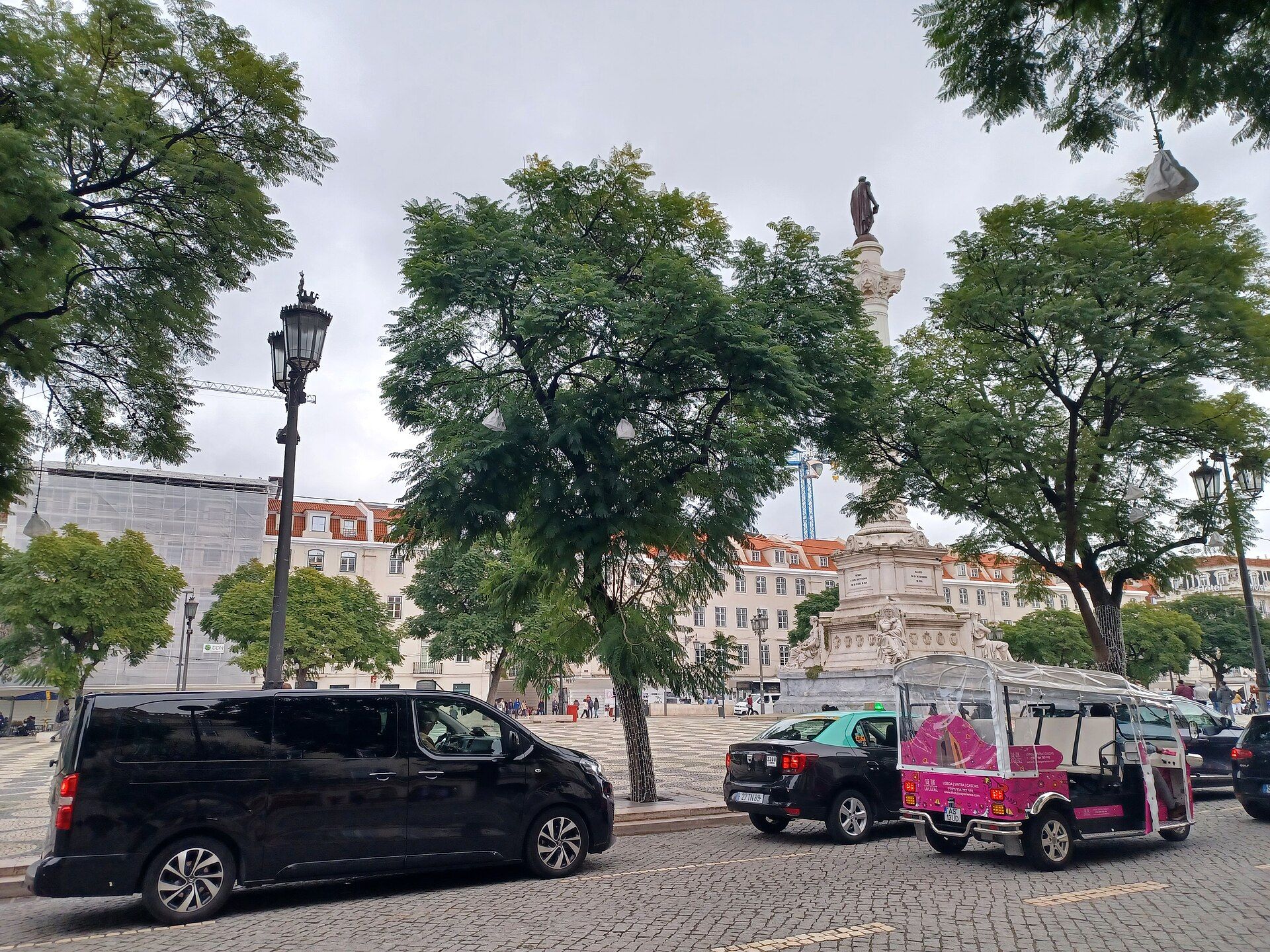 Praça do Rossio, em Lisboa, com árvores, a coluna de D. Pedro IV ao fundo e carros a passar